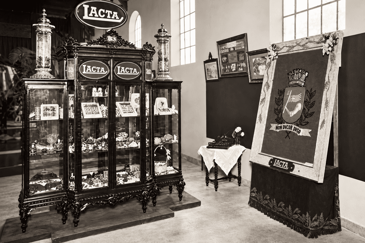 Vitrine antiga da Lacta com chocolates e caixas de bombons em exposição, ao lado de um estandarte com o brasão da cidade de São Paulo. Fonte: Imagens divulgação.